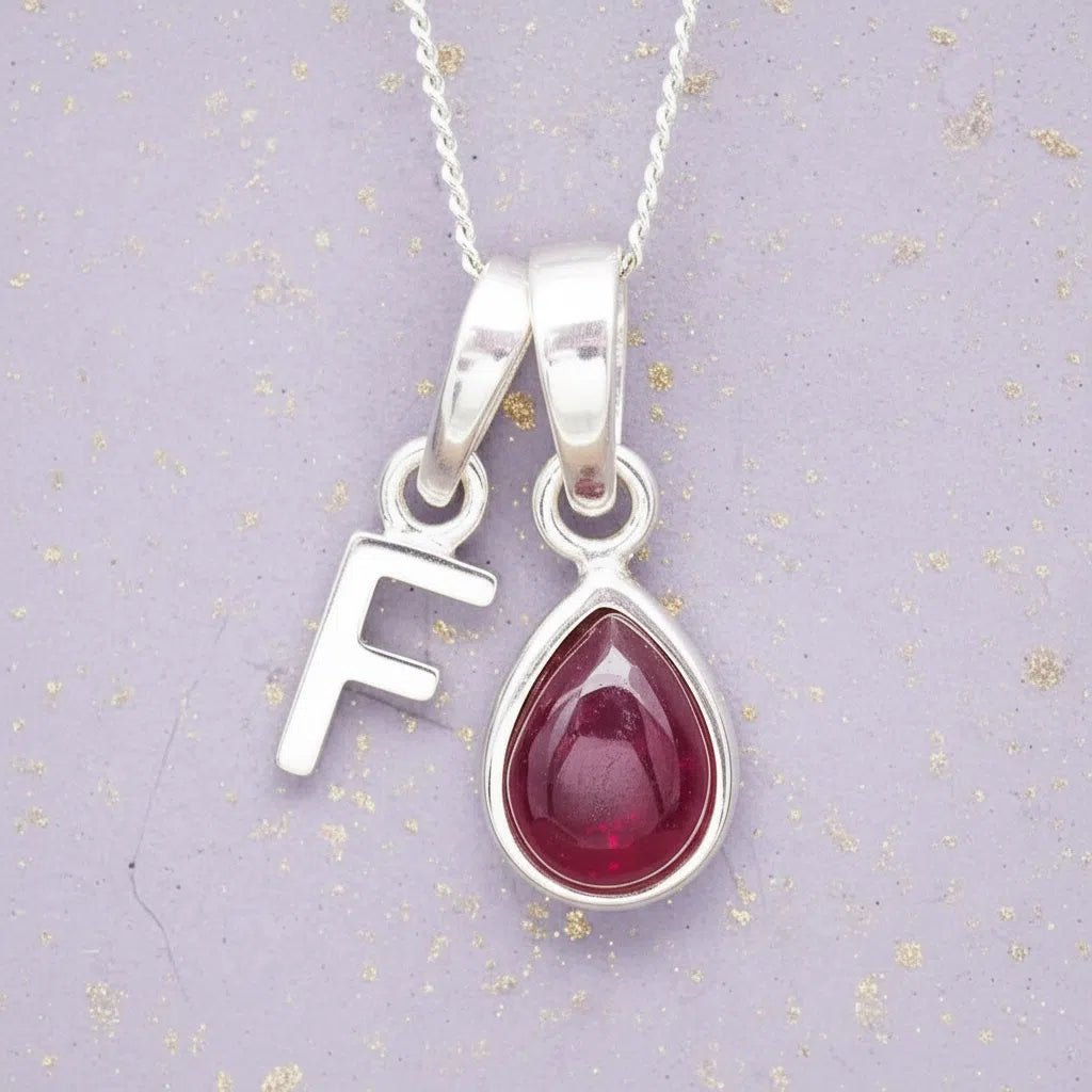 Sterling Silver necklace with 'F' initial pendant and July Birthstone Ruby gemstone pear-shaped pendant on a grey background.