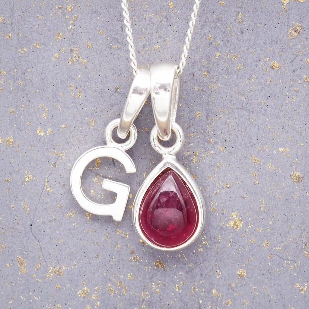 Sterling Silver necklace with 'G' initial pendant and July Birthstone Ruby gemstone pear-shaped pendant on a grey background.