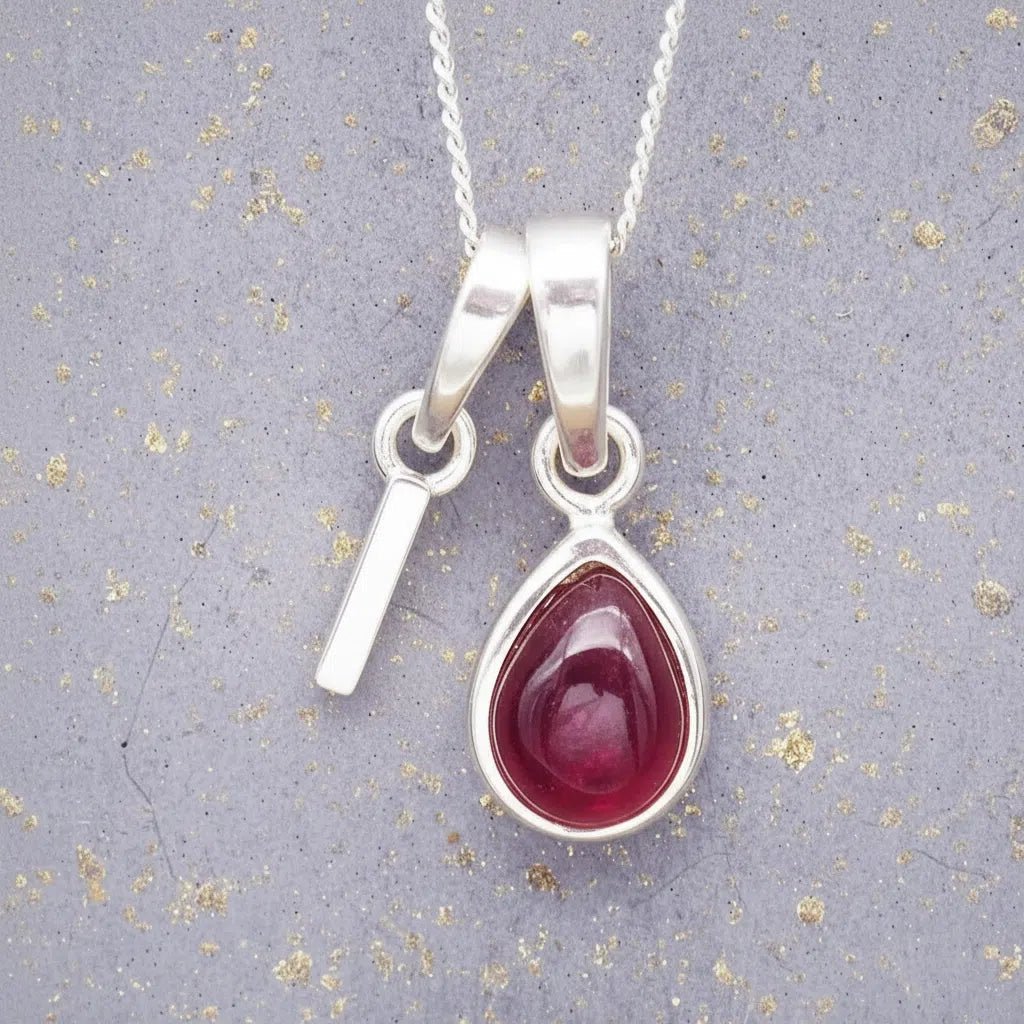 Sterling Silver necklace with 'I' initial pendant and July Birthstone Ruby gemstone pear-shaped pendant on a grey background.
