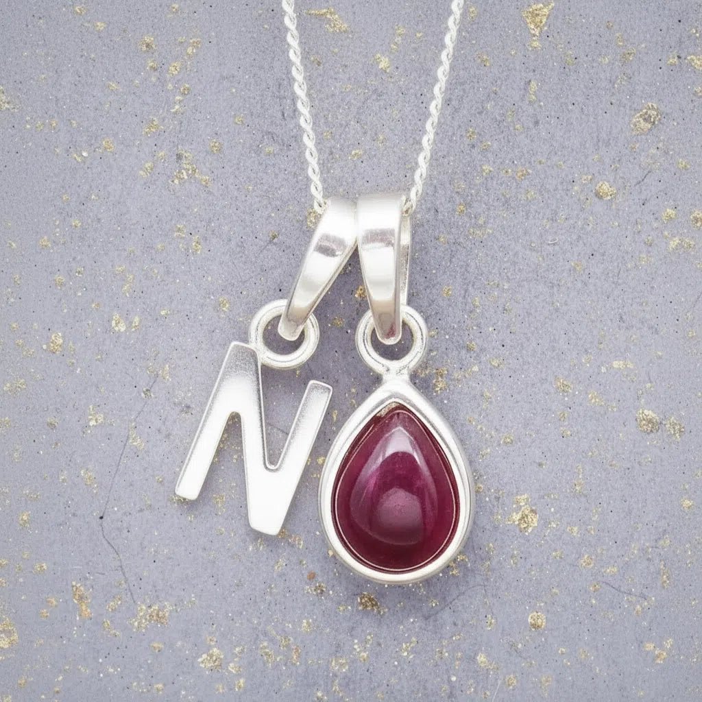 Sterling Silver necklace with 'N' initial pendant and July Birthstone Ruby gemstone pear-shaped pendant on a grey background.