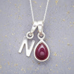 Sterling Silver necklace with 'N' initial pendant and July Birthstone Ruby gemstone pear-shaped pendant on a grey background.