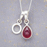 Sterling Silver necklace with 'O' initial pendant and July Birthstone Ruby gemstone pear-shaped pendant on a grey background.