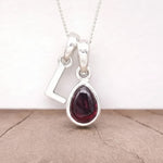 Silver Initial L pendant with a red teardrop-shaped Garnet gemstone on a beige surface.
