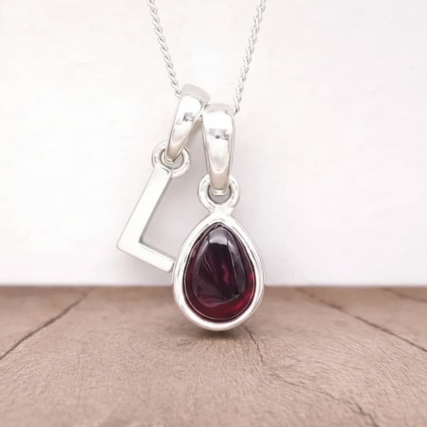 Silver Initial L pendant with a red teardrop-shaped Garnet gemstone on a beige surface.