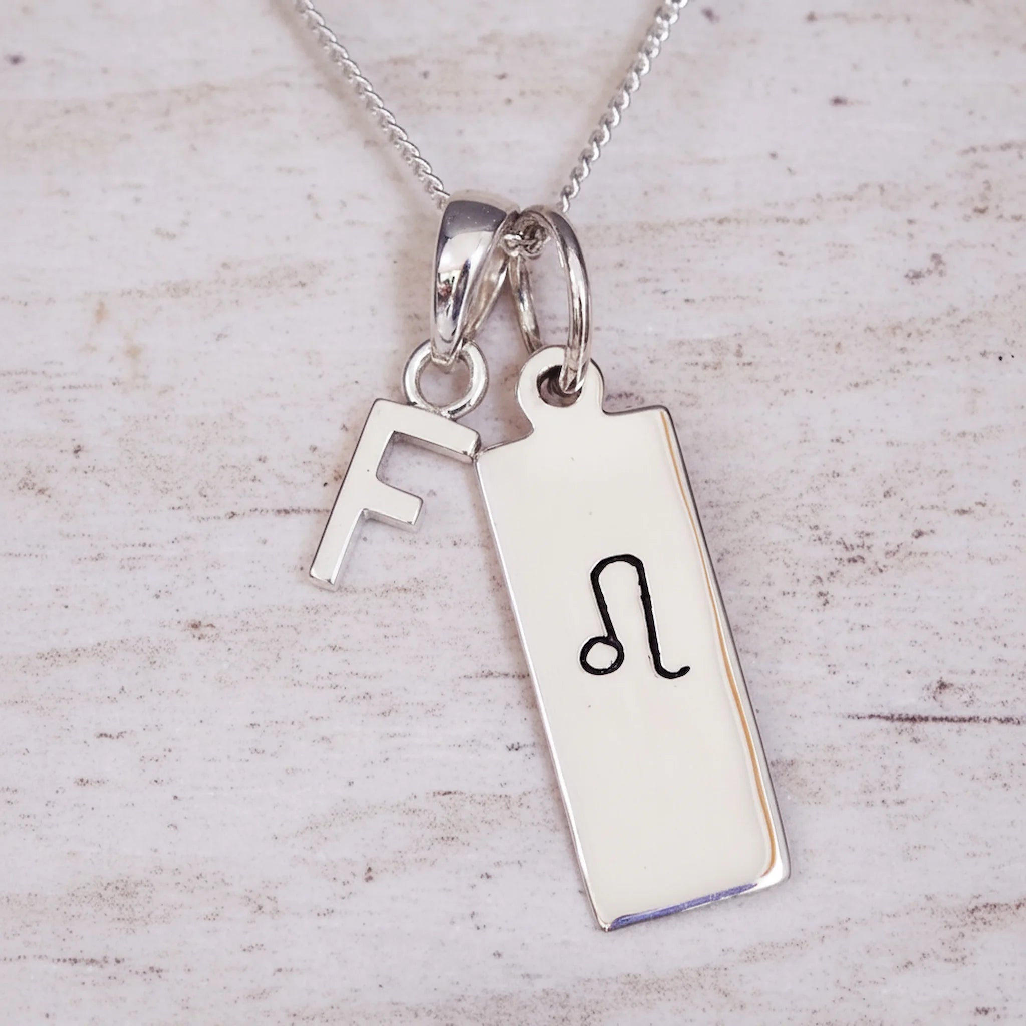 Sterling Silver necklace with a Leo Zodiac Star Sign pendant and a initial 'F' letter pendant on a light wooden background.
