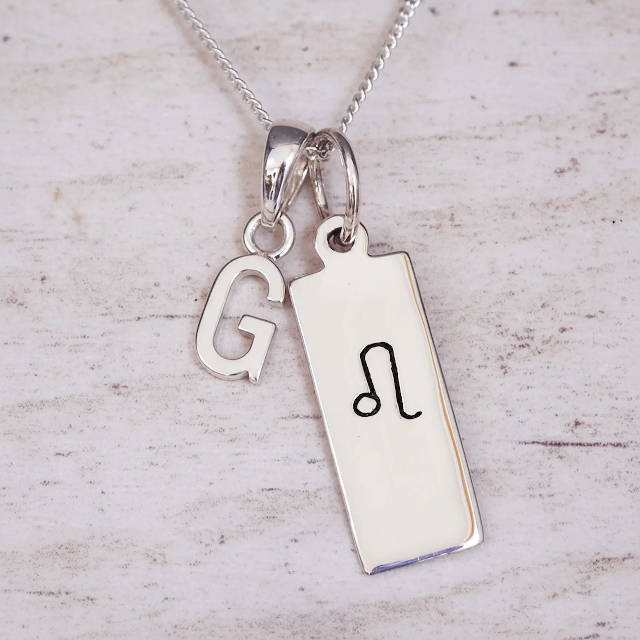 Sterling Silver necklace with a Leo Zodiac Star Sign pendant and a initial 'G' letter pendant on a light wooden background.