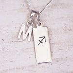 Sterling silver initial M and Sagittarius zodiac bar necklace, close-up of the letter charm and pendant on whitewashed wood