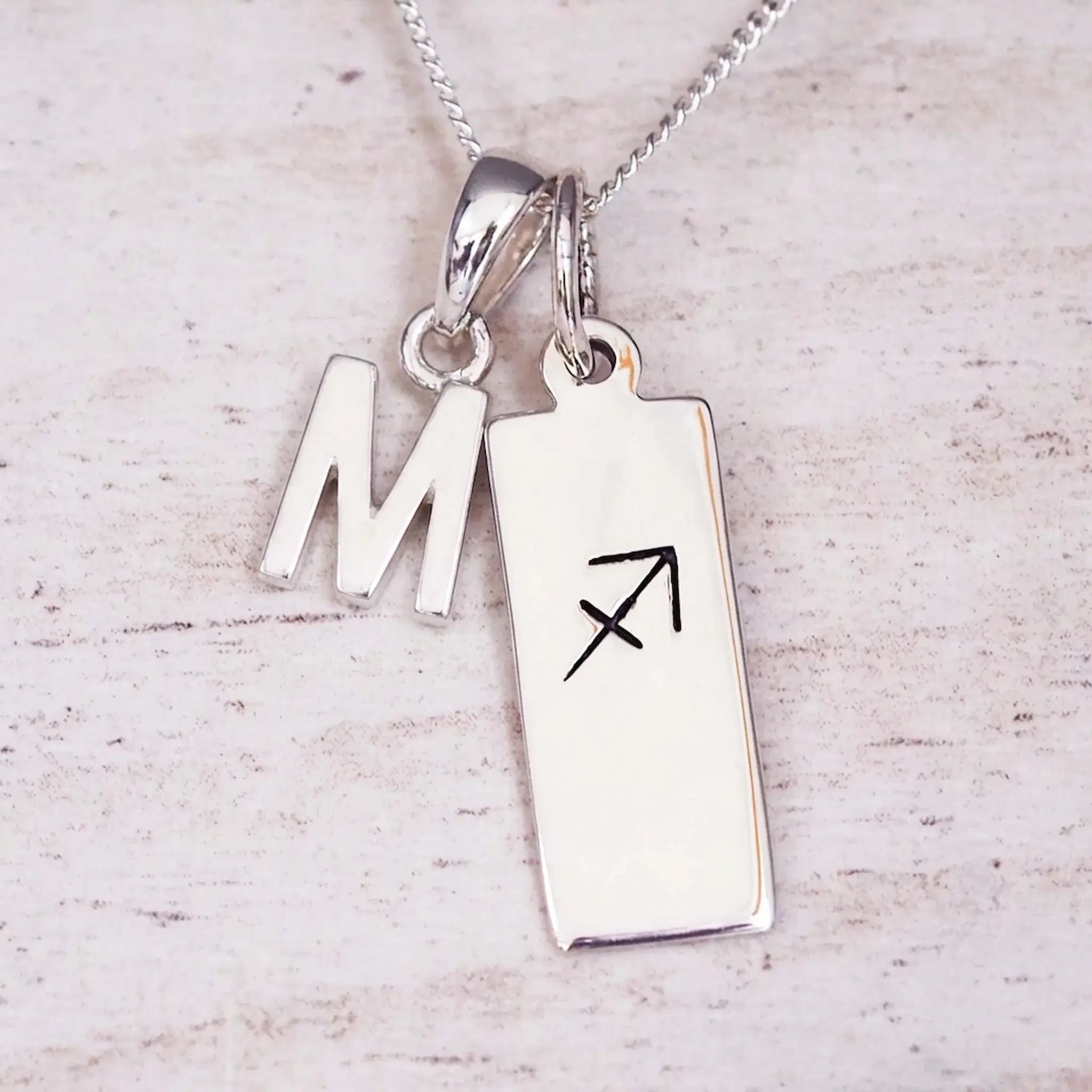 Sterling silver initial M and Sagittarius zodiac bar necklace, close-up of the letter charm and pendant on whitewashed wood