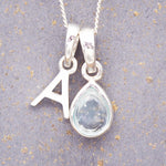 Silver november birthstone necklace with a pear-shaped topaz gemstone pendant and letter 'A' charm on a grey background.