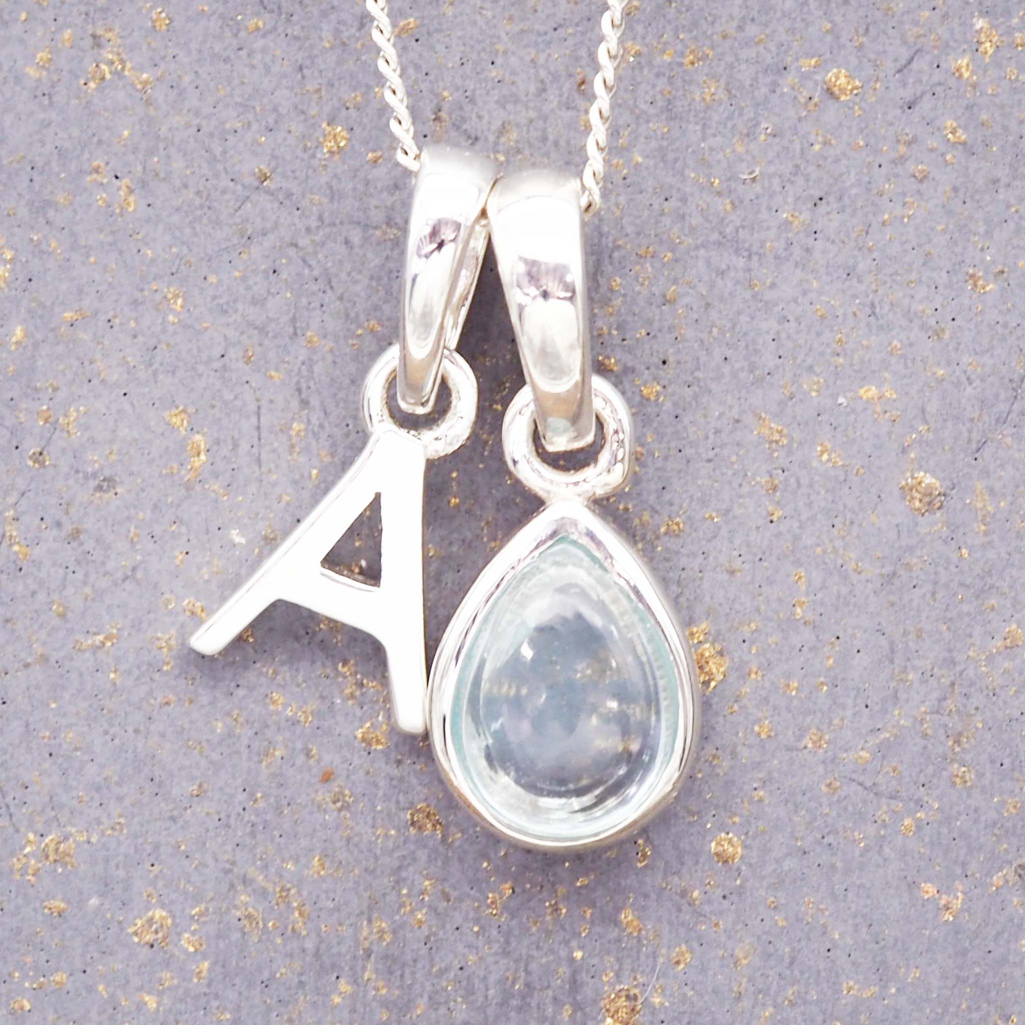 Silver november birthstone necklace with a pear-shaped topaz gemstone pendant and letter 'A' charm on a grey background.