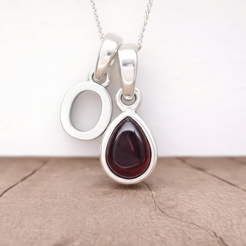 Silver Initial O necklace with a teardrop-shaped Garnet gemstone pendant hanging above a rustic wooden surface