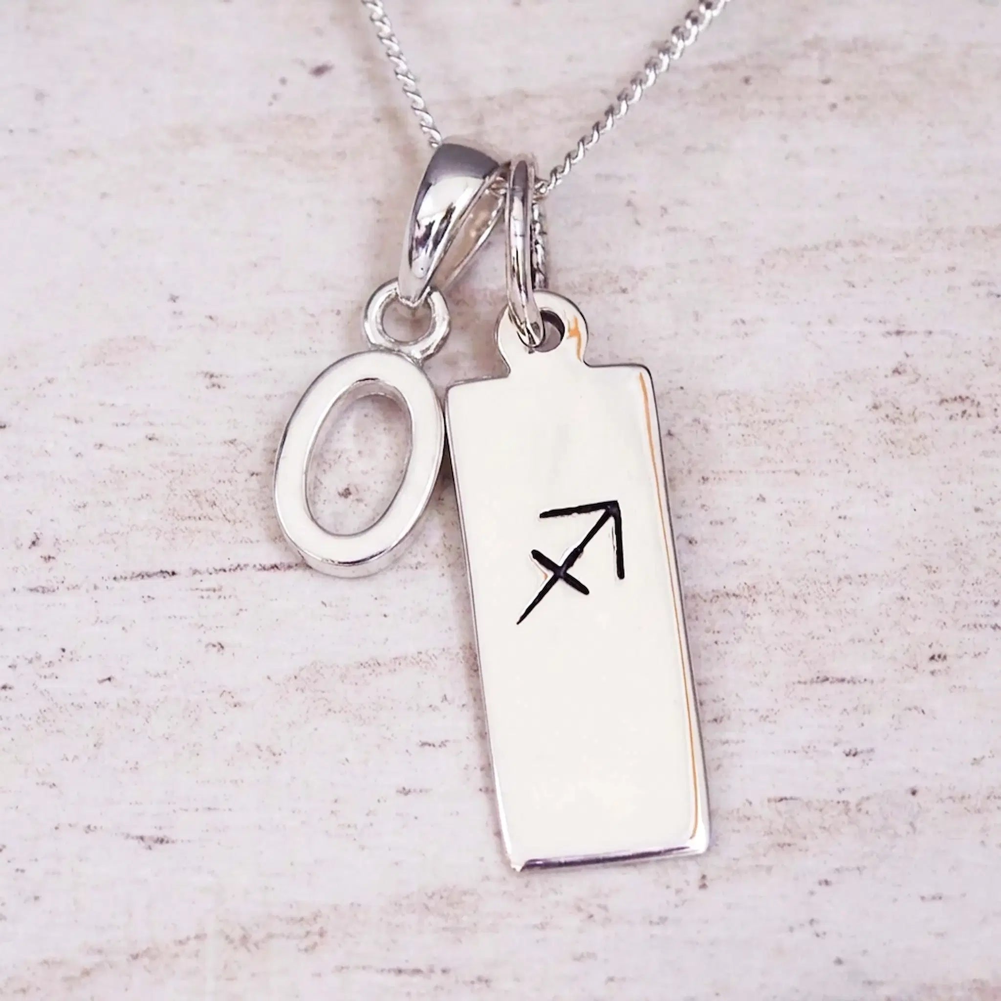 Sterling silver initial O and Sagittarius zodiac bar necklace, close-up of the letter charm and pendant on whitewashed wood