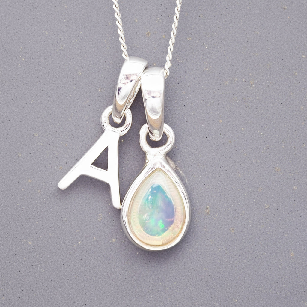 Silver october birthstone necklace with a pear-shaped opal pendant and a 'A' charm on a grey background.