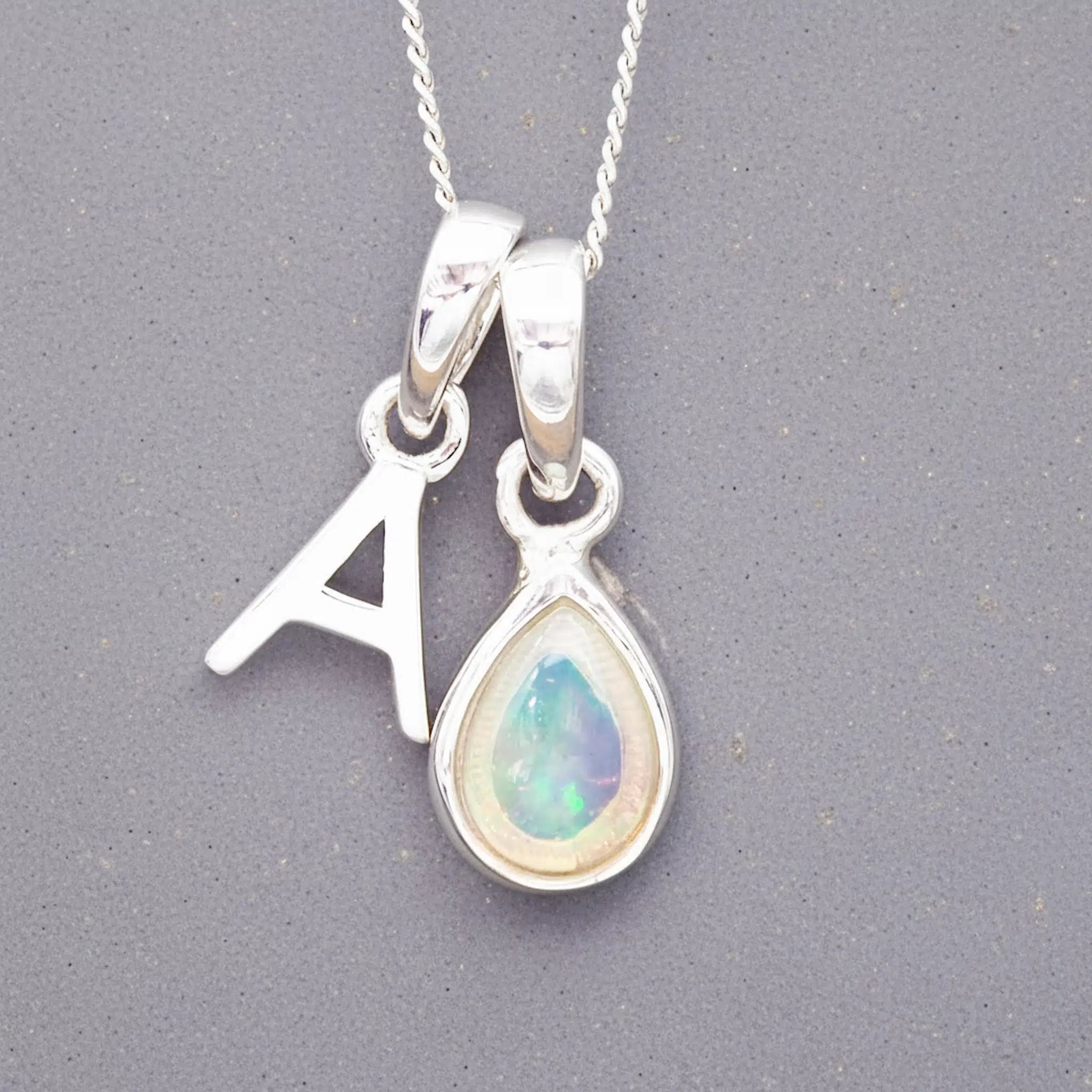 Silver october birthstone necklace with a pear-shaped opal pendant and a 'A' charm on a grey background.