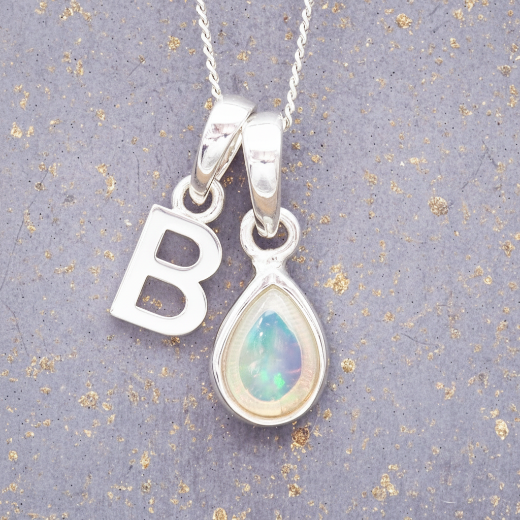 Silver necklace with 'B' pendant and teardrop-shaped opal pendant on a textured gray background.
