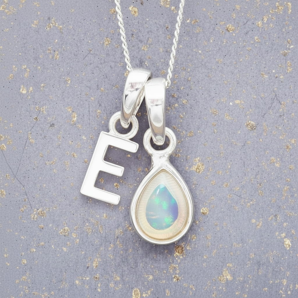 Silver necklace with 'E' pendant and teardrop-shaped opal pendant on a textured gray background.