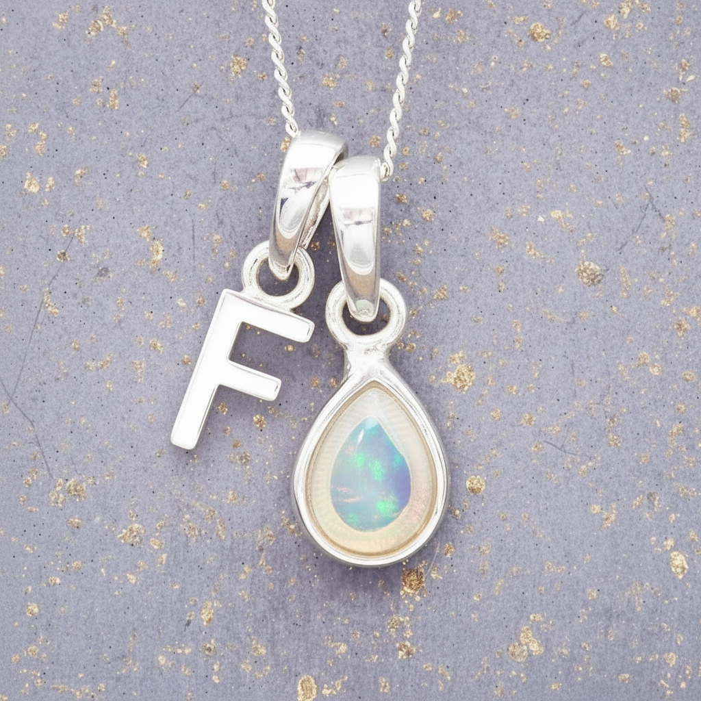 Silver necklace with a teardrop-shaped opal pendant and letter 'F' charm on a textured gray background.
