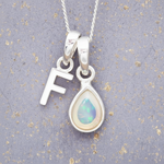 Silver necklace with a teardrop-shaped opal pendant and letter 'F' charm on a textured gray background.