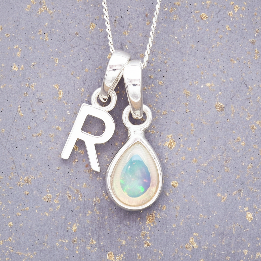 Silver necklace with 'R' pendant and teardrop-shaped opal pendant on a textured grey background.