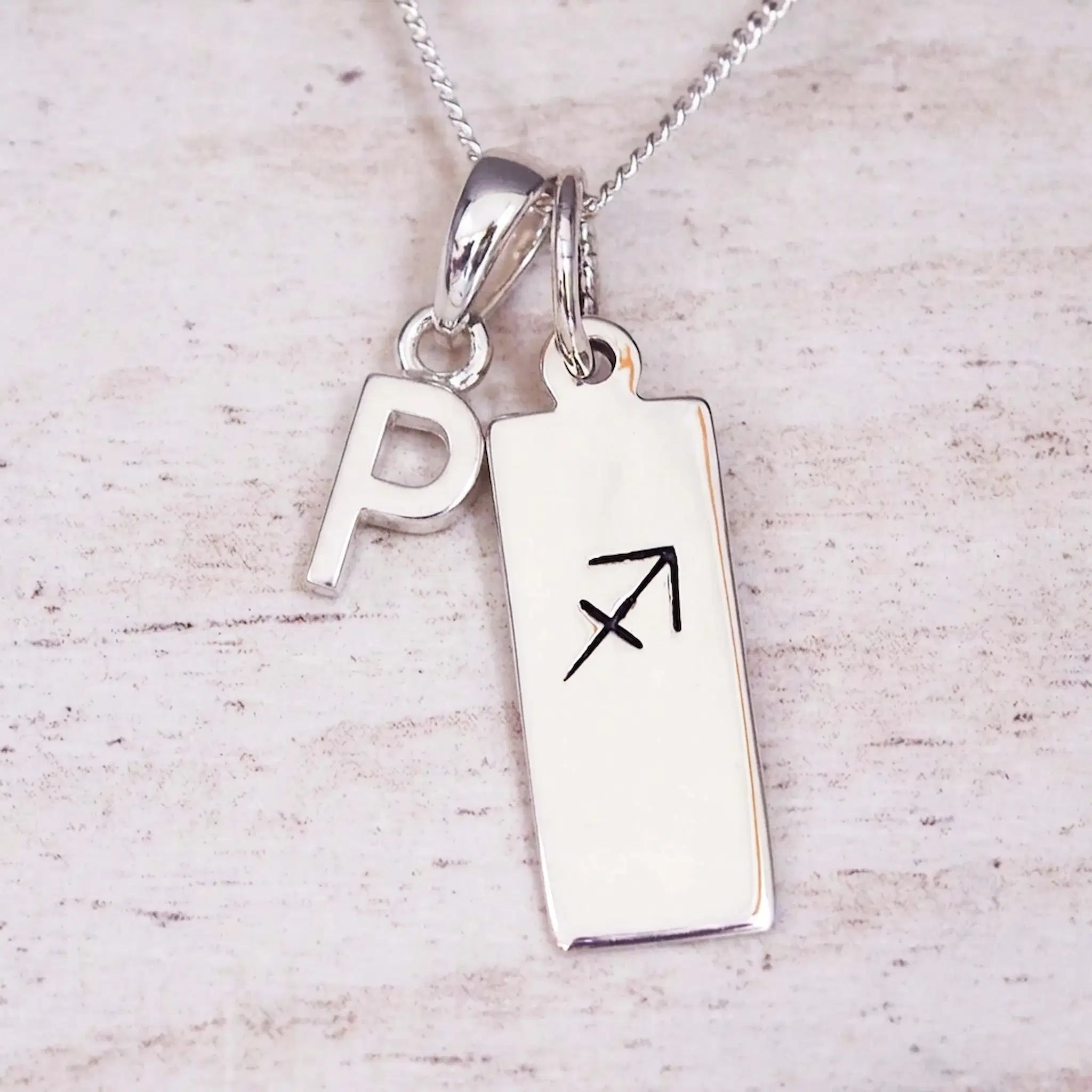 Sterling silver initial P and Sagittarius zodiac bar necklace, close-up of the letter charm and pendant on whitewashed wood