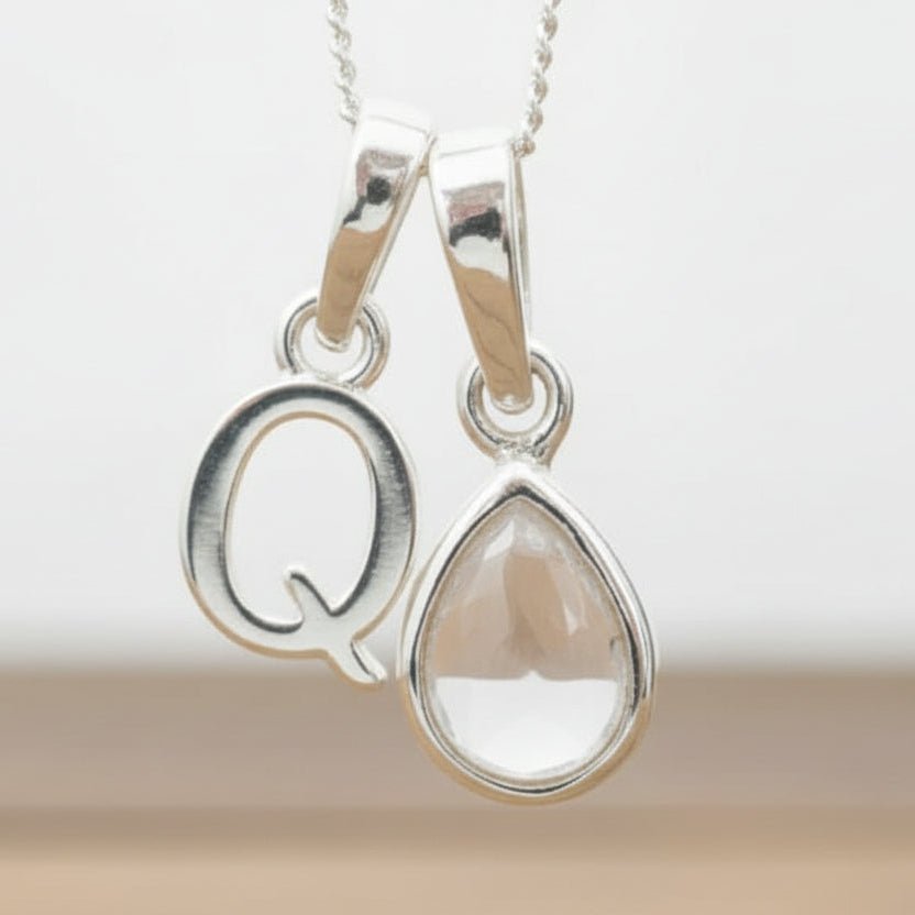 Silver Initial Q necklace with a teardrop-shaped Herkimer Quartz crystal pendant hanging above a rustic wooden surface