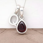 Silver Initial Q necklace with a teardrop-shaped Garnet gemstone pendant hanging above a rustic wooden surface