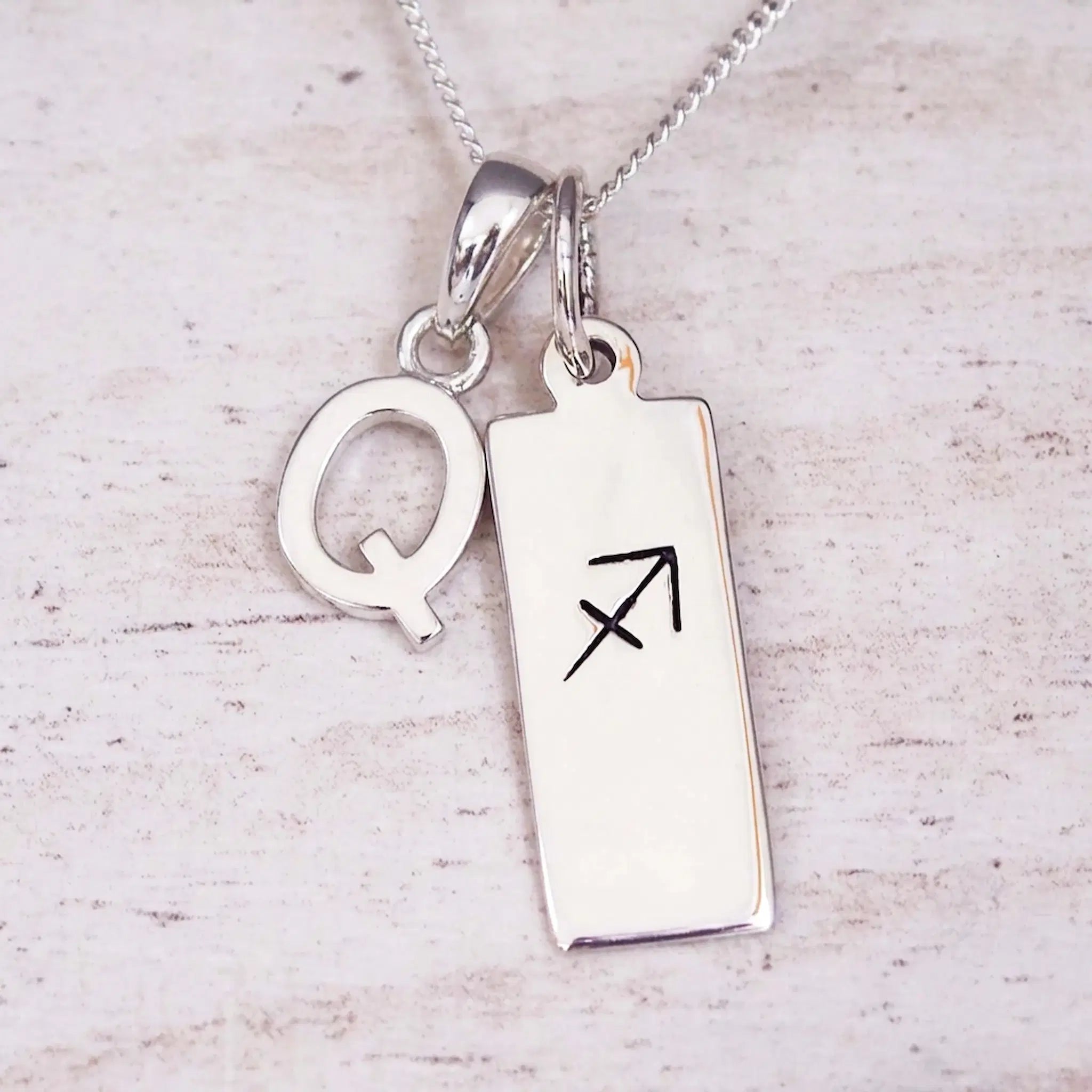 Sterling silver initial Q and Sagittarius zodiac bar necklace, close-up of the letter charm and pendant on whitewashed wood