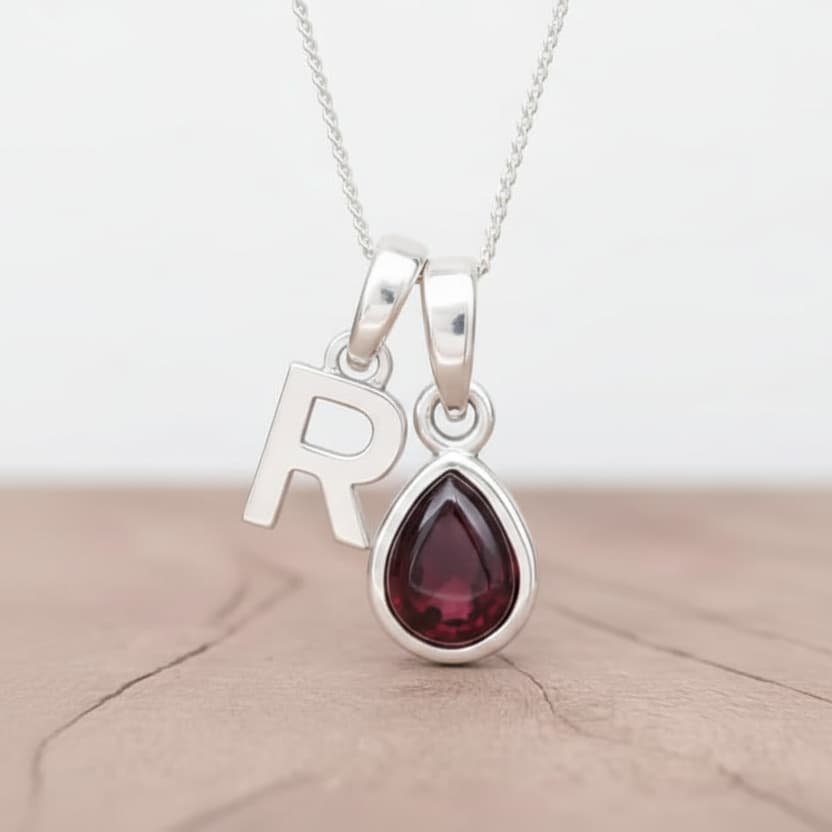 Silver necklace with a teardrop-shaped garnet gemstone pendant and letter 'R' charm on a light wooden surface.