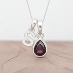 Silver Initial S necklace with a teardrop-shaped Garnet gemstone pendant hanging above a rustic wooden surface