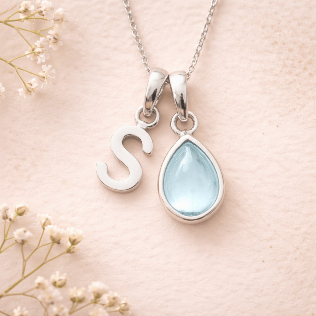 Silver necklace with 'S' pendant and teardrop-shaped light blue stone on a soft pink background with white flowers.
