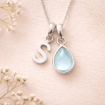 Silver necklace with 'S' pendant and teardrop-shaped light blue stone on a soft pink background with white flowers.