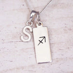 Sterling silver initial S and Sagittarius zodiac bar necklace, close-up of the letter charm and pendant on whitewashed wood