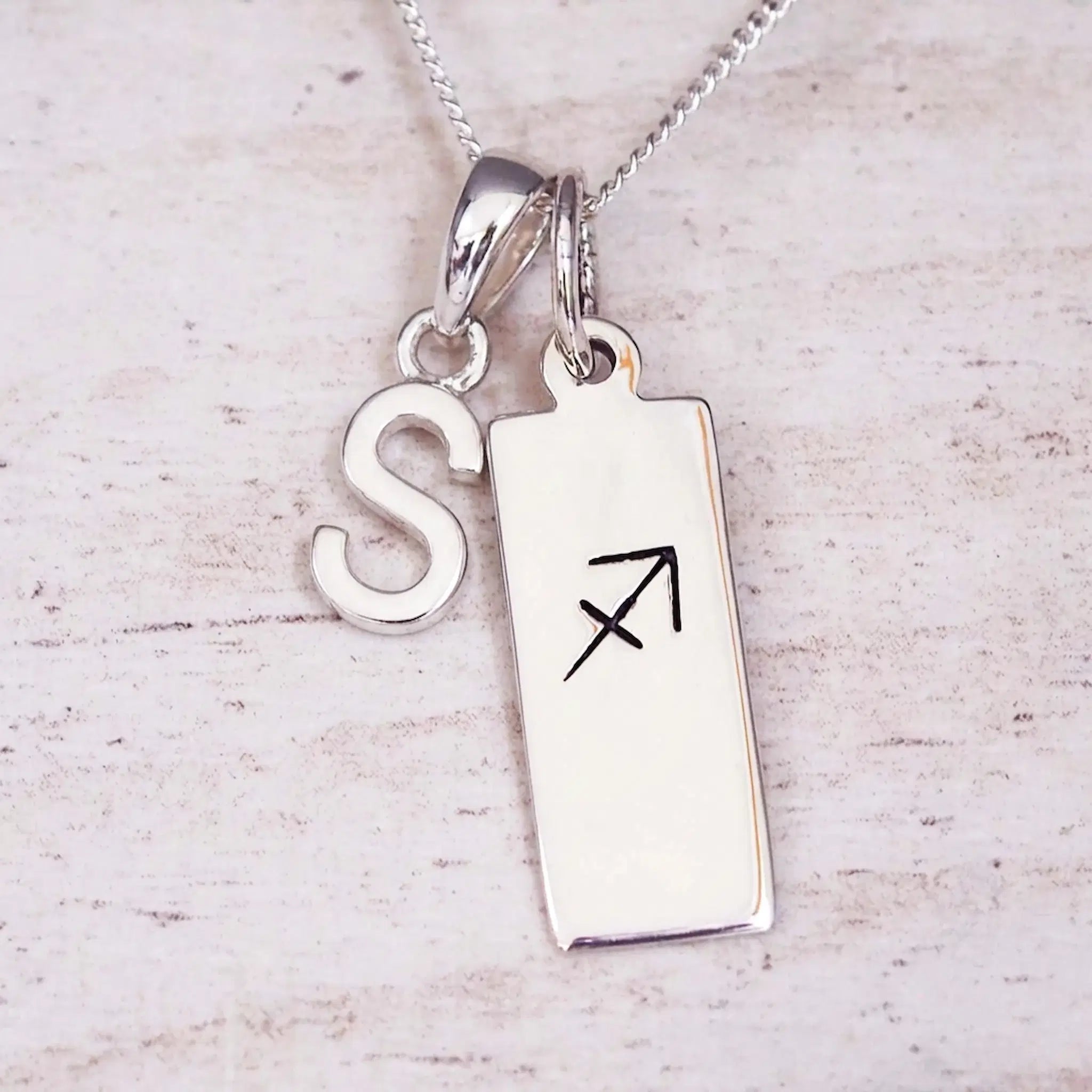 Sterling silver initial S and Sagittarius zodiac bar necklace, close-up of the letter charm and pendant on whitewashed wood