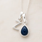 Two silver pendants on a chain against a light background, a uppercase Z pendant and a pear shaped september birthstone sapphire pendant.