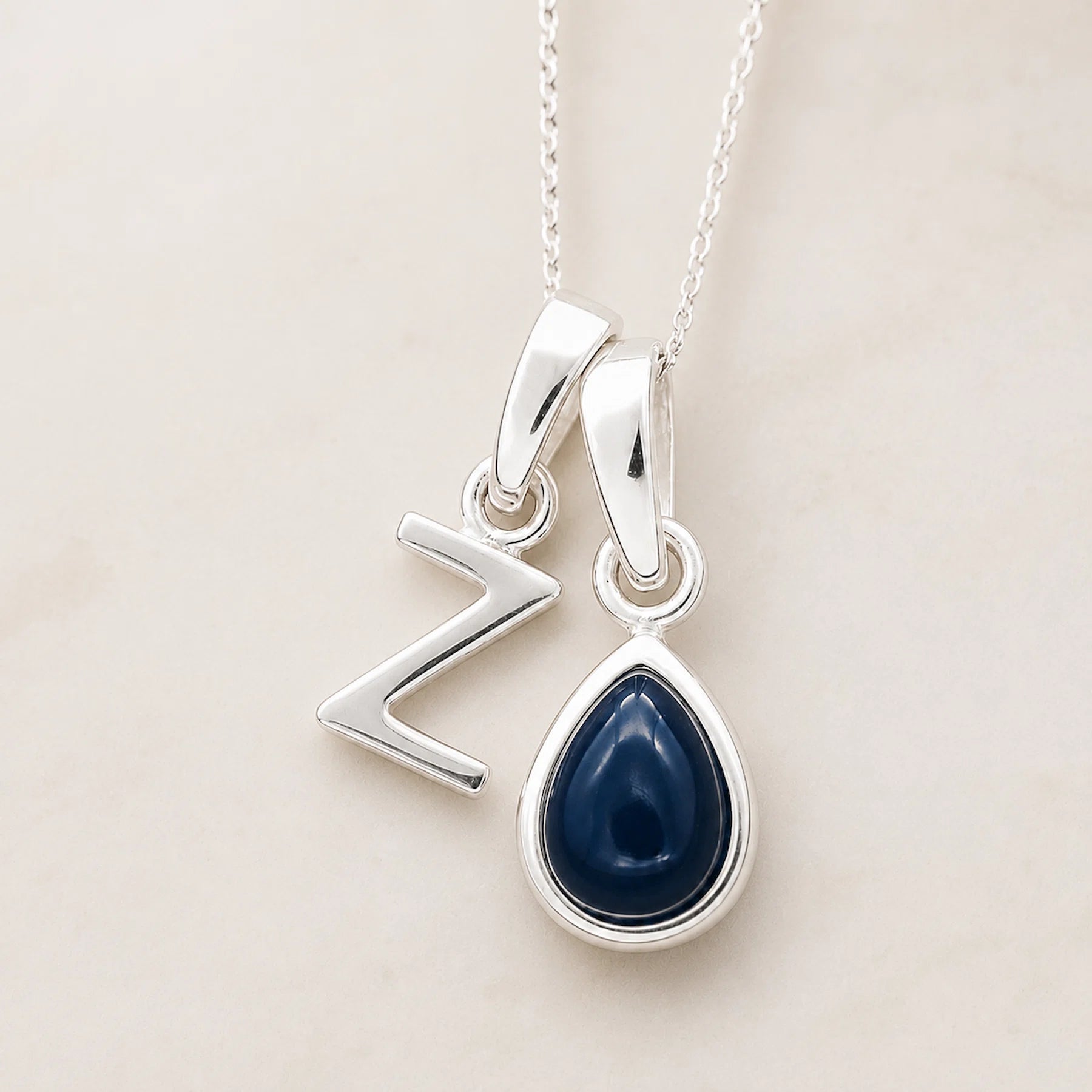 Two silver pendants on a chain against a light background, a uppercase Z pendant and a pear shaped september birthstone sapphire pendant.
