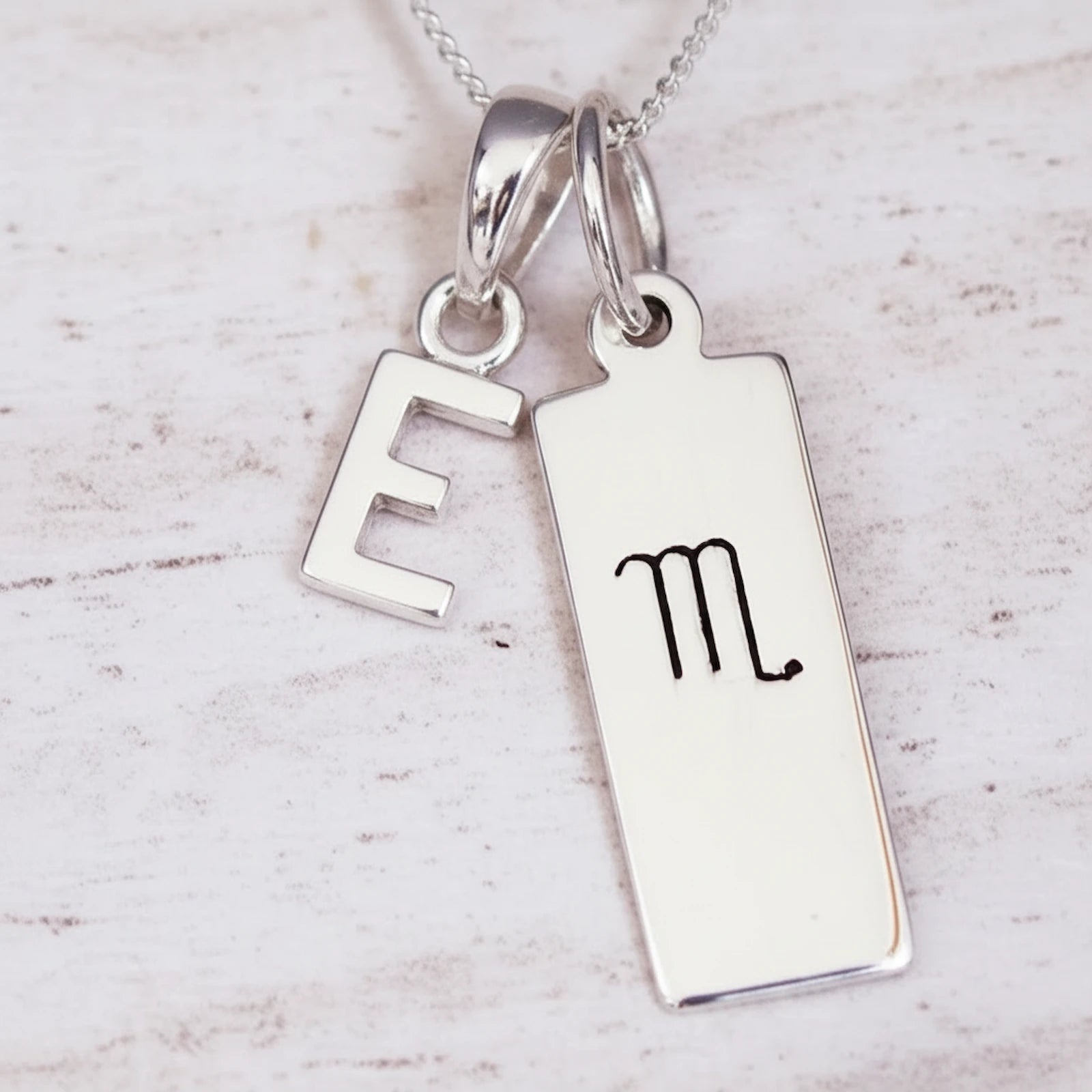 Sterling Silver necklace with 'E' initial pendant and hand drawn Scorpio zodiac symbol pendant on a light wooden background