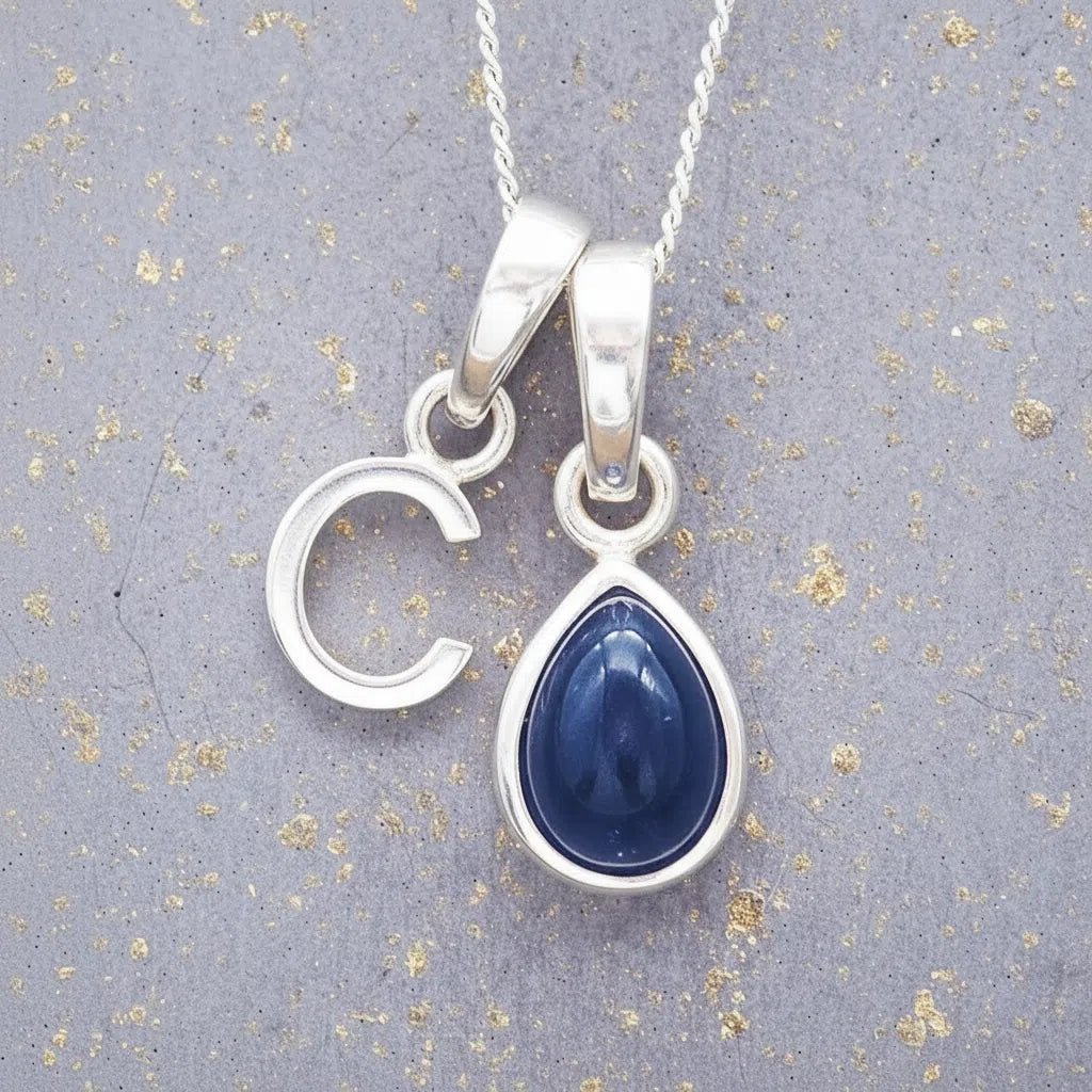 Sterling silver 'C' initial necklace with a September birthstone blue sapphire pear-shaped pendant on a grey background.