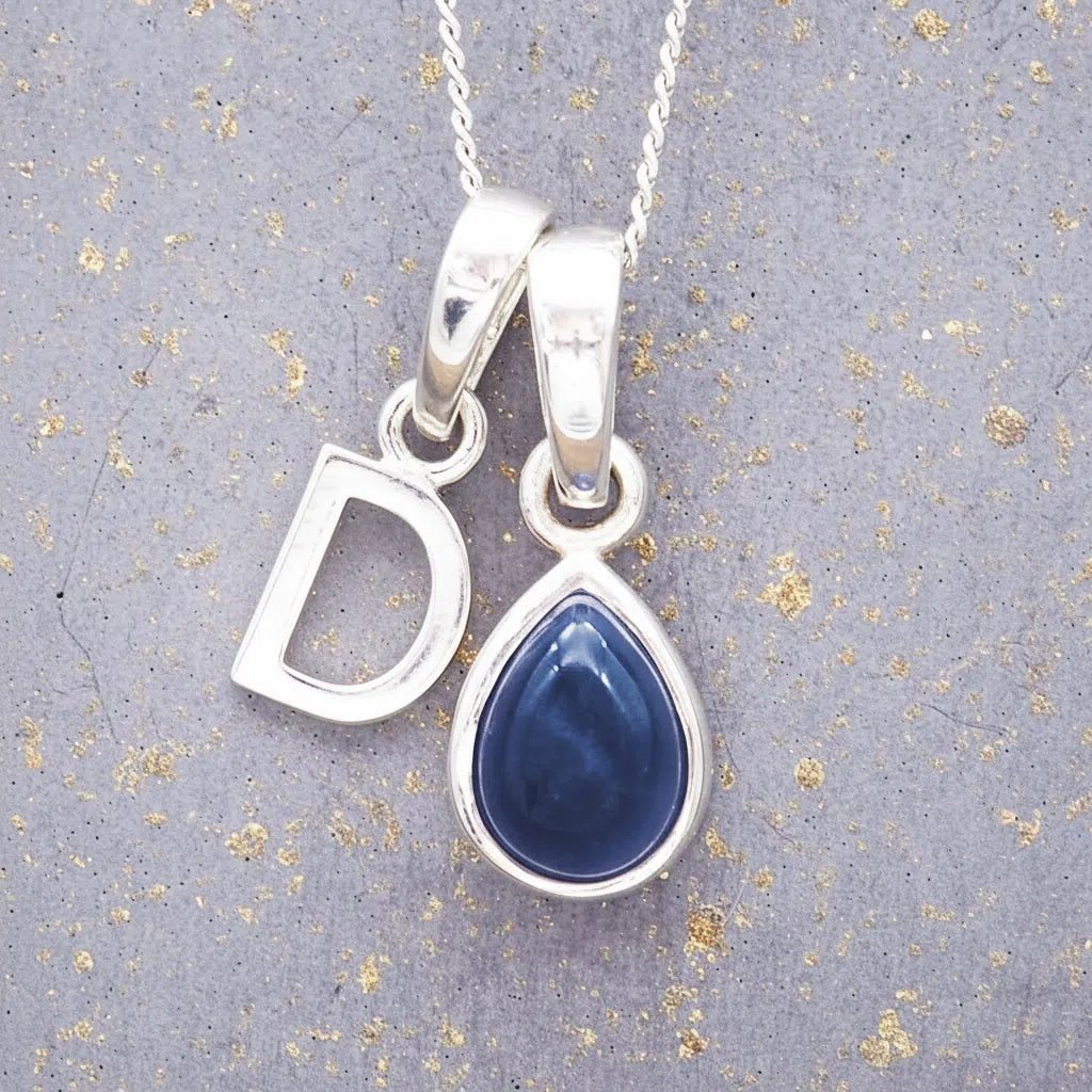 Sterling silver 'D' initial necklace with a September birthstone blue sapphire pear-shaped pendant on a grey background.