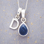 Sterling silver 'D' initial necklace with a September birthstone blue sapphire pear-shaped pendant on a grey background.