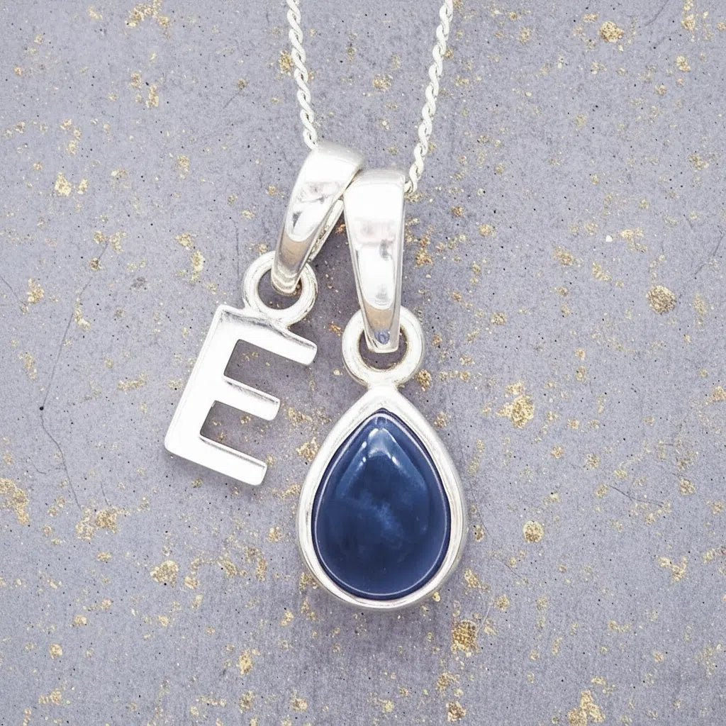 Sterling silver 'E' initial necklace with a September birthstone blue sapphire pear-shaped pendant on a grey background.
