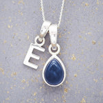 Sterling silver 'E' initial necklace with a September birthstone blue sapphire pear-shaped pendant on a grey background.