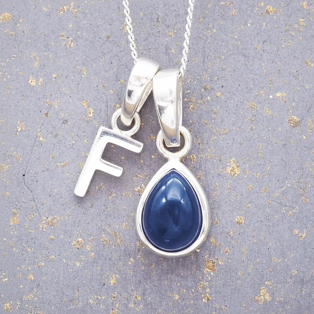 Sterling silver 'F' initial necklace with a September birthstone blue sapphire pear-shaped pendant on a grey background.