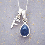Sterling silver 'F' initial necklace with a September birthstone blue sapphire pear-shaped pendant on a grey background.