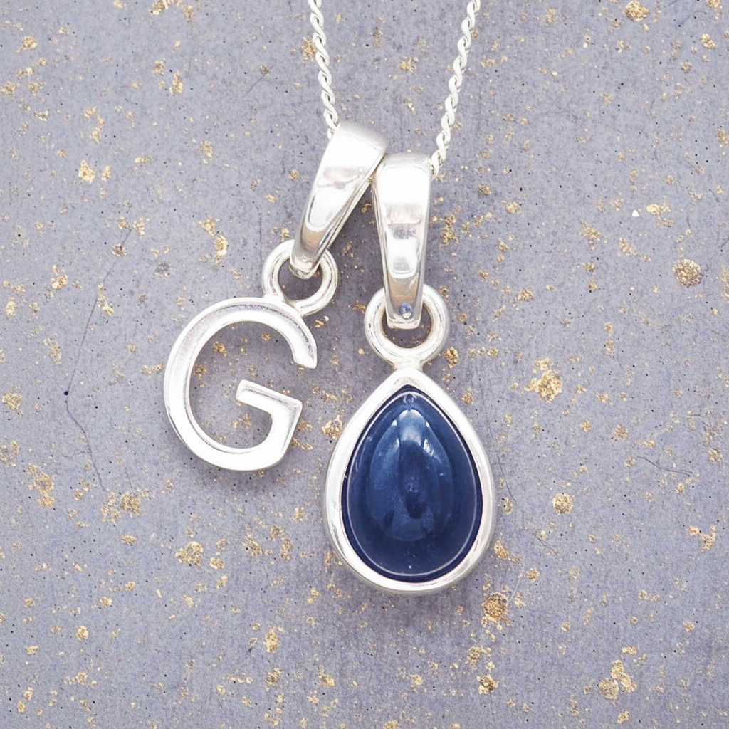 Sterling Silver necklace with a 'G' initial pendant with a blue teardrop-shaped september birthstone sapphire gemstone on a textured grey background
