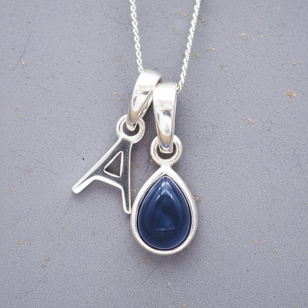 Sterling silver 'A' initial necklace with a September birthstone blue sapphire pear-shaped pendant on a grey background.