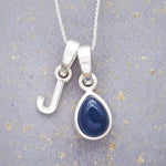 Sterling silver 'J' initial necklace with a September birthstone blue sapphire pear-shaped pendant on a grey background.