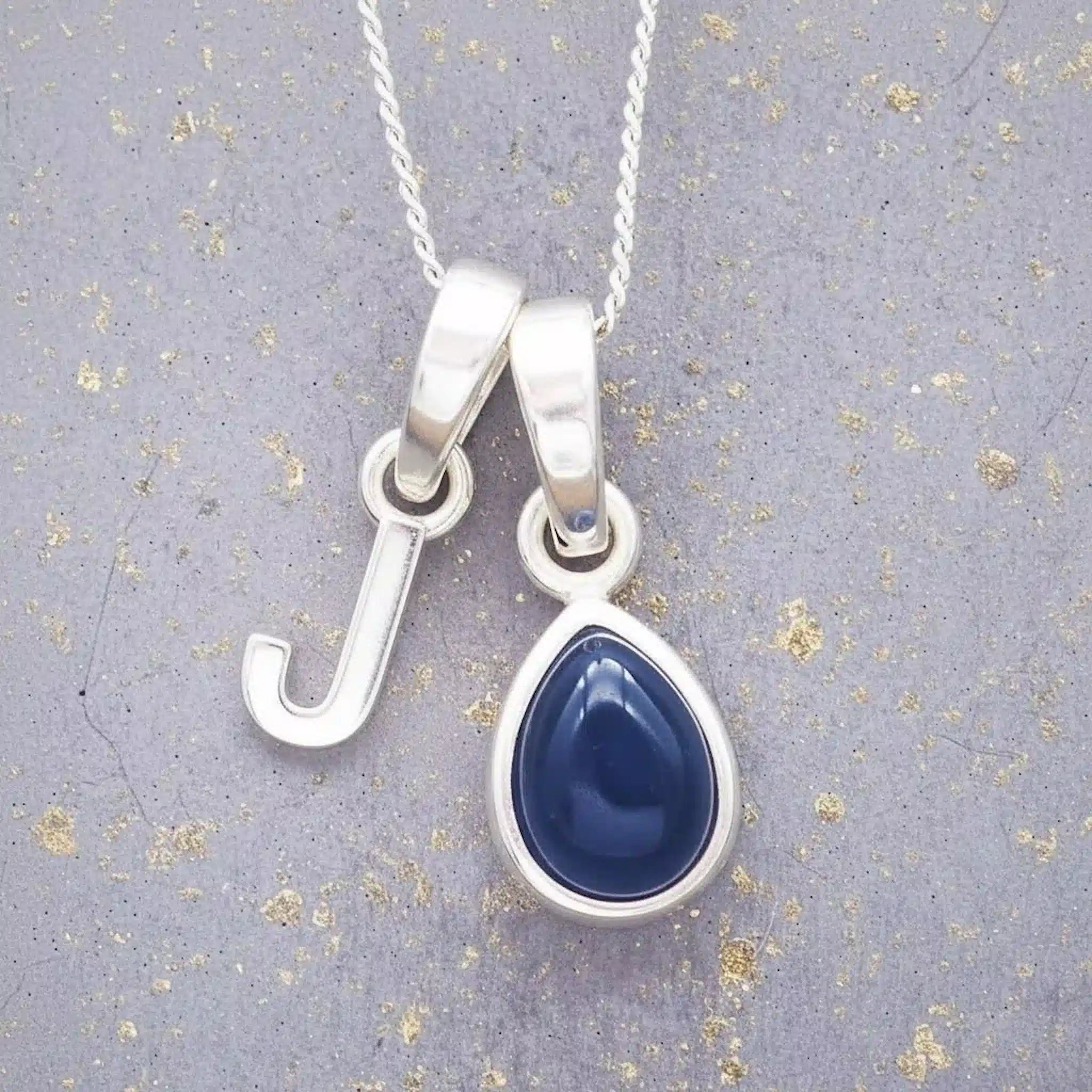 Sterling silver 'J' initial necklace with a September birthstone blue sapphire pear-shaped pendant on a grey background.