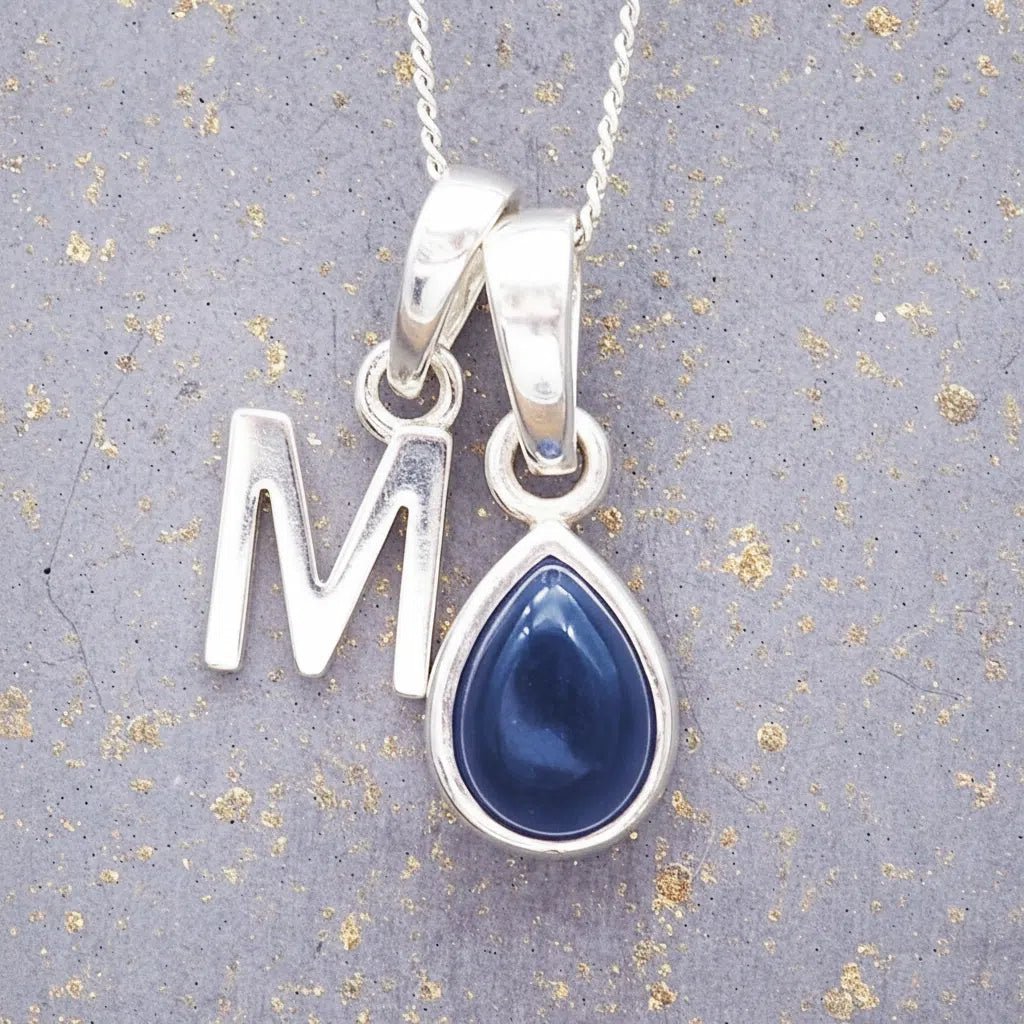 Sterling silver 'M' initial necklace with a September birthstone blue sapphire pear-shaped pendant on a grey background.