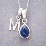 Sterling silver 'M' initial necklace with a September birthstone blue sapphire pear-shaped pendant on a grey background.