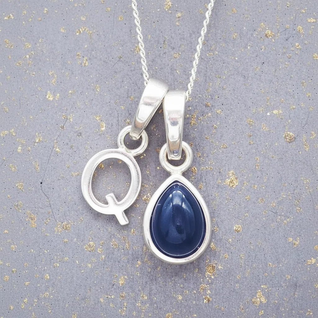 Sterling silver 'Q' initial necklace with a September birthstone blue sapphire pear-shaped pendant on a grey background.
